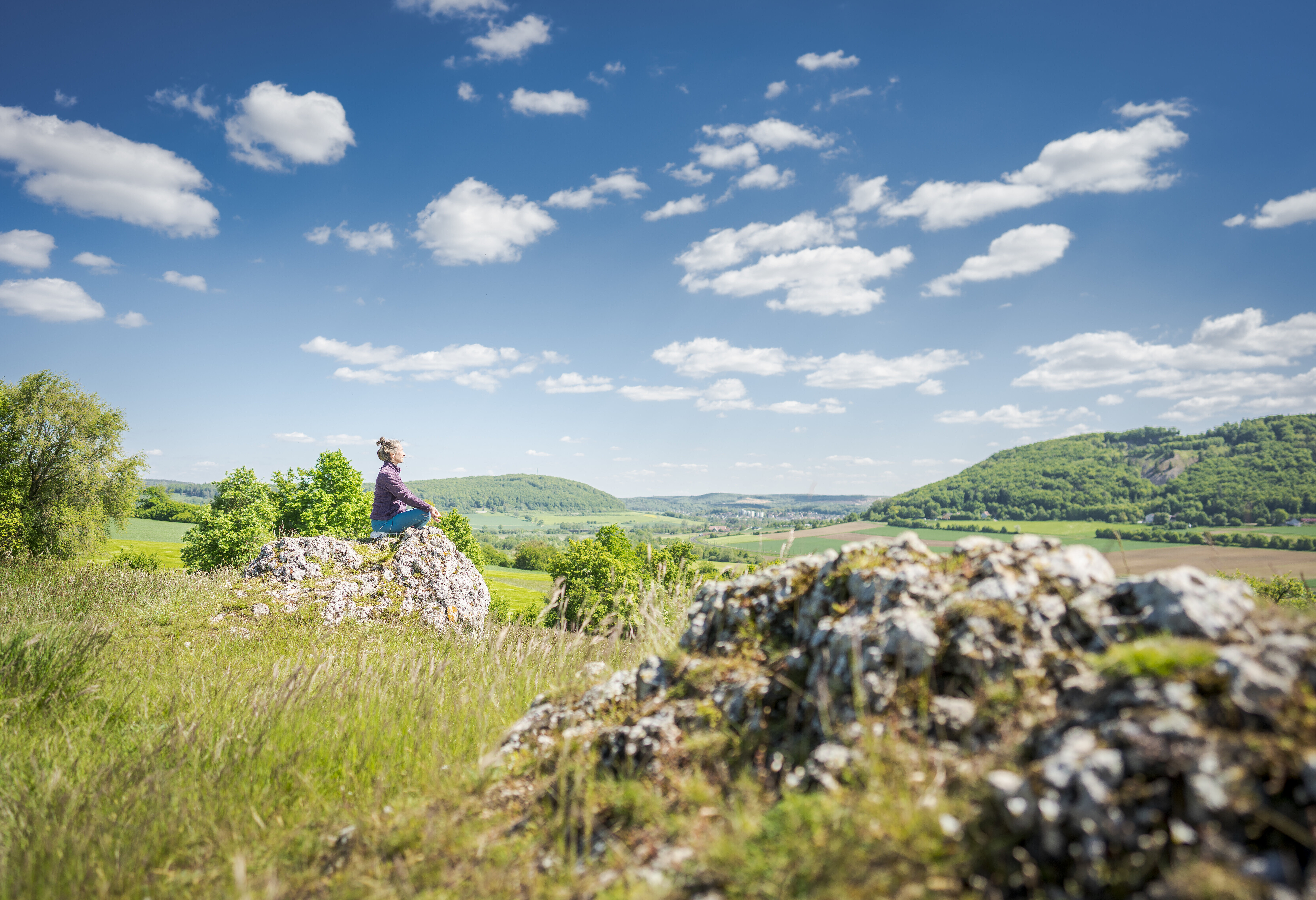 Junge Frau auf einem Felsblock im Altmühltal in Yogaposition, blauer Himmel, weiße Wolken, Sonnenschein